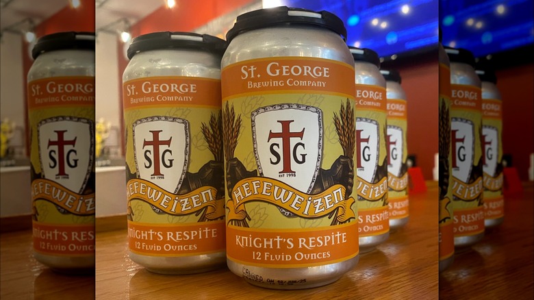 4 cans on a table, the design features an orange and yellow color with a central shield logo with a red cross and the letters SG est 1998. Behind the shield, knight's gauntlets hold sheaves of wheat. The can reads: St. George Brewing Company, Hefeweizen, Knight's respite 12 fluid ounces.