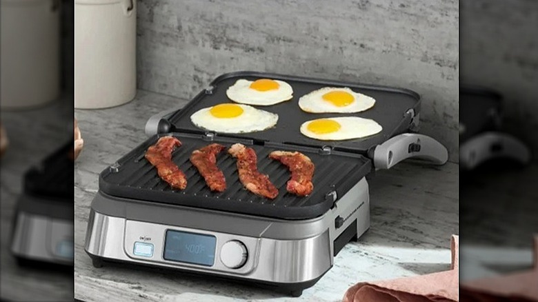 Cuisinart electric griddle with four pieces of bacon on one side and four sunny side up eggs on the other side cooking.