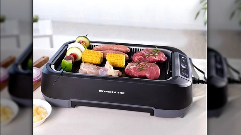 Ovente griddle & grill appliance cooking steaks, sausages, and vegetables on countertop.