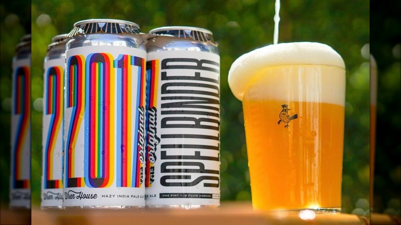 A glass of hazy golden beer with a thick foamy head sitting beside a four-pack of Wren House Brewing Co.'s Spellbinder cans with colorful, retro-style lettering.