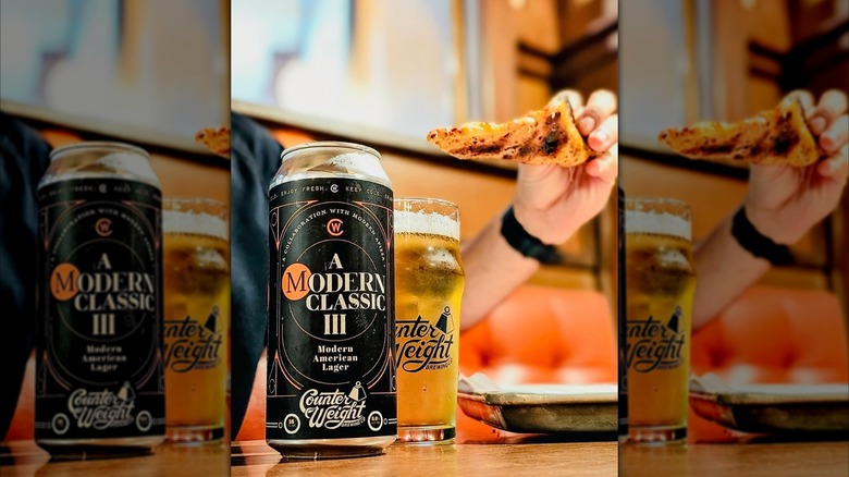 A can and glass of Counter Weight Brewing's Modern Classic lager on a table while someone holds a slice of pizza in the background.