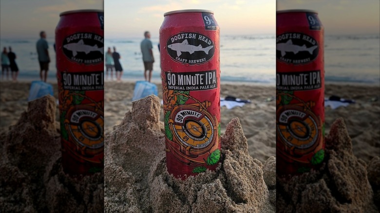 A can of Dogfish Head 90 Minute IPA sitting in sand on the beach at sunset, with the ocean and people in the distance.