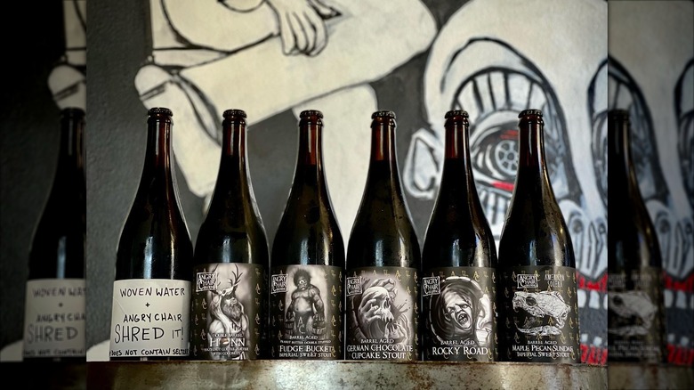 Six bottles of Angry Chair barrel-aged stouts lined up on a metal surface with a graffiti-style mural background.