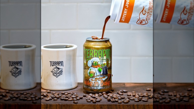 A can of Terrapin Wake-n-Bake Coffee Oatmeal Imperial Stout and a Terrapin mug sitting on a wooden surface surrounded by coffee beans while coffee from a Jittery Joe's cup is being poured in the can.