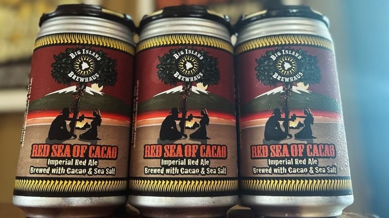 Three cans of Big Island Brewhouse Red Sea of Cacao Imperial Red Ale, and a blurred taproom background.