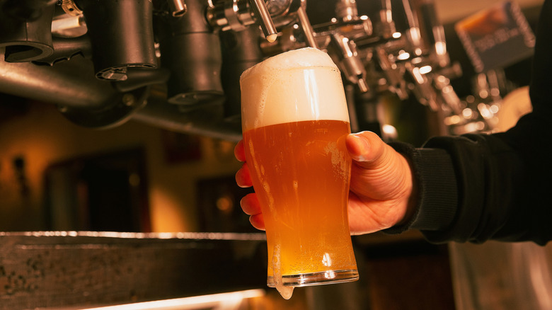 A freshly poured pint of hazy beer with a frothy head held under draft taps inside a bar