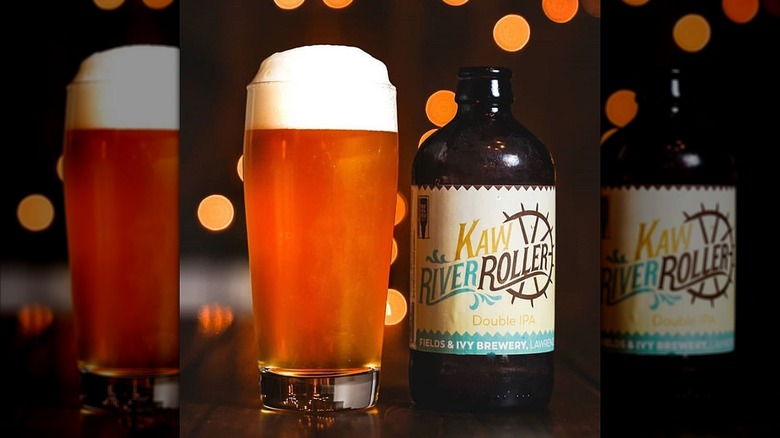 A glass of hazy golden beer with a thick foam head sitting beside a short, wide bottle of Fields & Ivy Brewery's Kaw River Roller Double IPA.