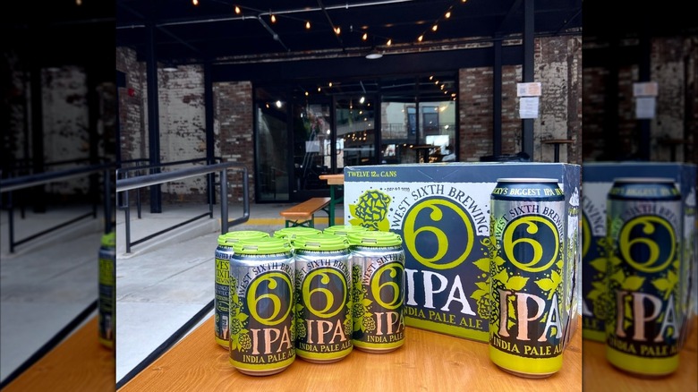 A twelve-pack and several cans of West Sixth IPA sitting on an outdoor brewery patio table, with string lights and an industrial taproom in the background.