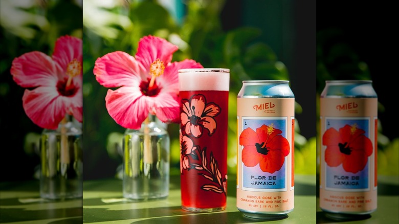 A can and glass of deep red hibiscus beer from Miel Brewery sit beside a fresh pink hibiscus flower in a glass vase set outdoors.