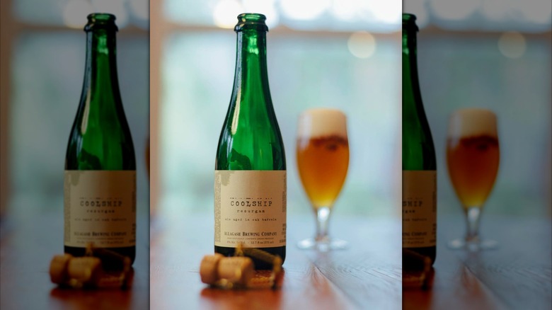 An uncorked green bottle of Allagash Coolship Resurgam sitting on a wooden table with a tulip glass of golden, foamy beer blurred in the background.