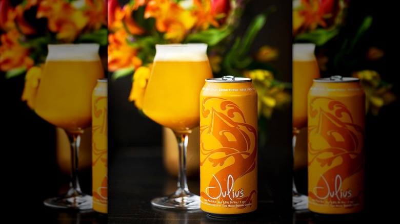 An orange can of Tree House Brewing's Julius IPA beside a stemmed glass filled with hazy, golden beer set in front of flowers