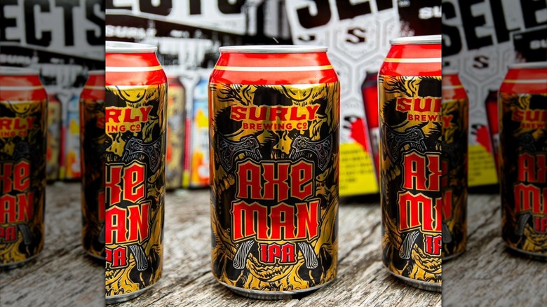 Three cans of Surly Brewing Co.'s Axe Man IPA with bold red and gold metallic artwork arranged on a wooden surface