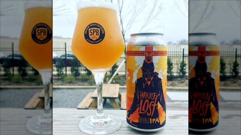 A tulip glass filled with hazy golden beer next to a can of Southern Prohibition Brewing's Paradise Lost Double IPA on an outdoor picnic table