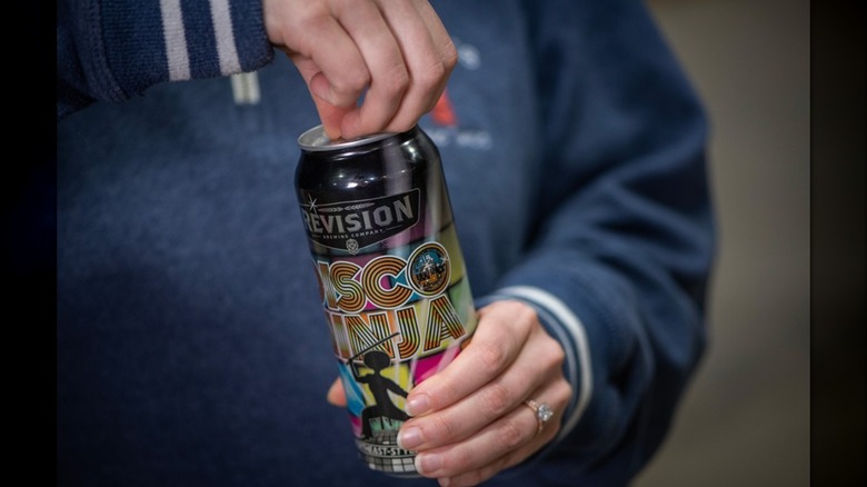 A person opening a can of Revision Brewing's Disco Ninja IPA, which features colorful disco-inspired artwork and a silhouette of a ninja dancing