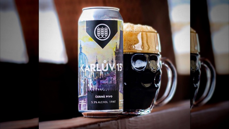 A pint can of Schilling Beer Co.'s Karlův 13° next to a dark, frothy mug of beer on a wooden beam