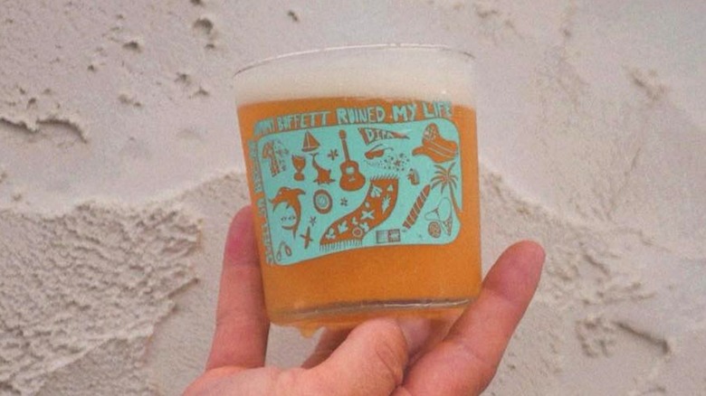 A glass of hazy beer with the logo for Jimmy Buffett Ruined My Life on it being held near sand.