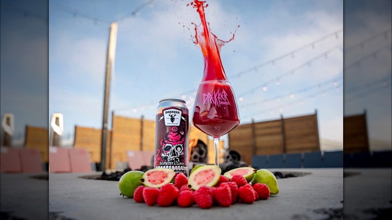 A can of Drekker's Braaaaaaaains Strawberry and Guava next to a glass overflowing with bright red liquid, surrounded by fresh raspberries and guava on a concrete fire pit