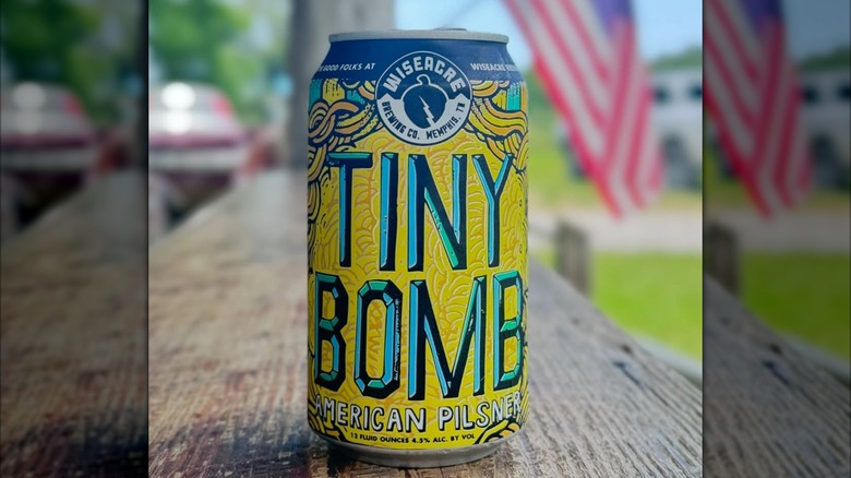 A can of Wiseacre Brewing's Tiny Bomb on a wooden surface, with a blurred American flag in the background