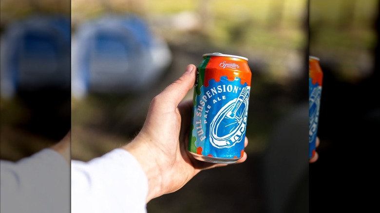 A person holding a can of Squatters Full Suspension Pale Ale with a blurred campground and tents in the background