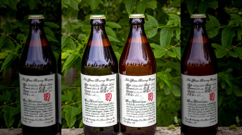 Two bottles of New Glarus Brewing's R&D series standing side by side outdoors, showcasing handwritten-style labels with green leaves blurred behind them