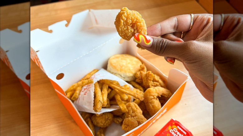 Woman holds up piece of fried shrimp from Popeyes Shrimp Tackle box with fried shrimp, fries, biscuit, next to packet of creole cocktail sauce
