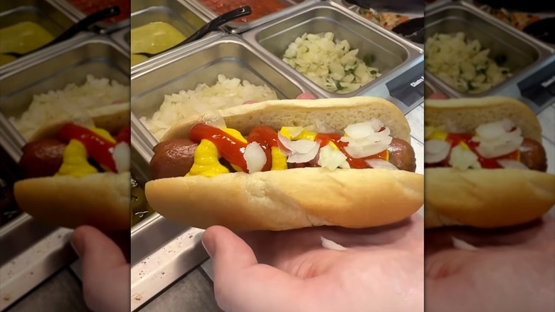 Hand holding ampm hot dog with ketchup, mustard, and onions in front of condiment tray