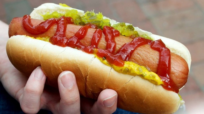 Hand holding hot dog with ketchup, mustard, and relish