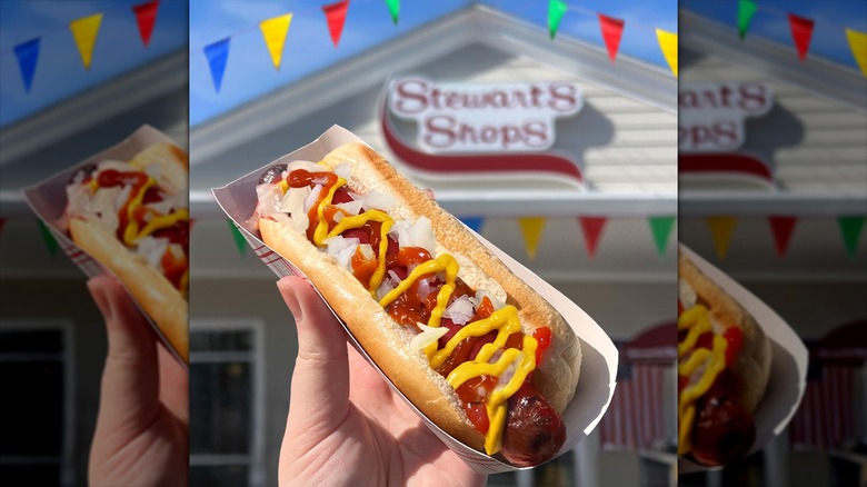 Hand holding up hot dog with several toppings in front of Stewart's Shops location