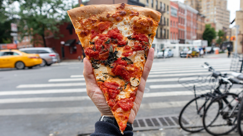 A close-up shot of a slice of veggie pizza held in one hand against a blurred backdrop of New York City