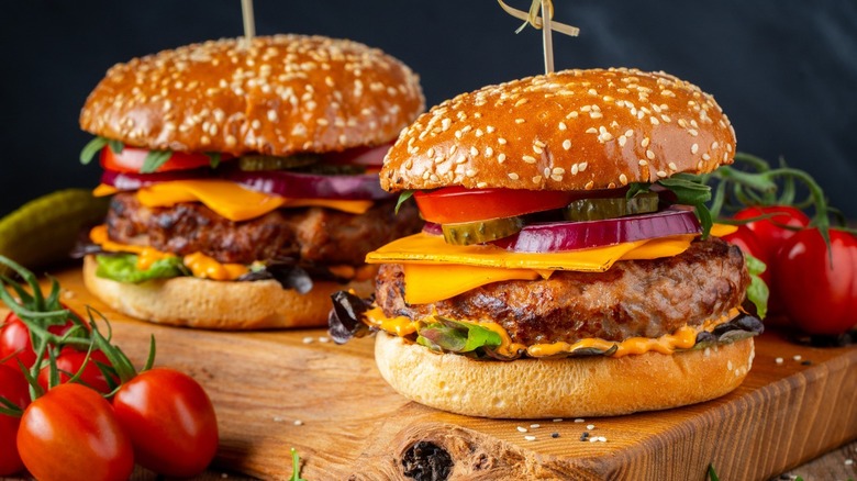 Two burgers with patty, cheese, veggies, over a wooden board 