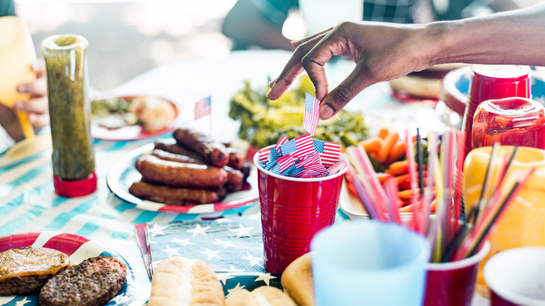 person taking miniature American flag from plastic cup on table surrounded by grilled meats and vegetables