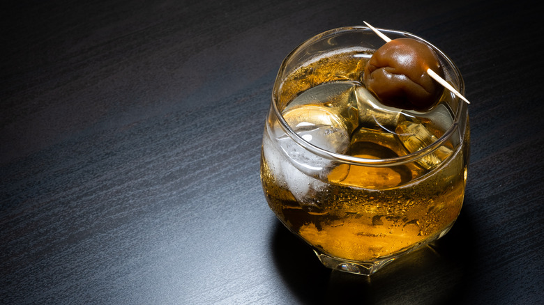 Glass of amber liquid with ice in it, an toothpick with an olive on top