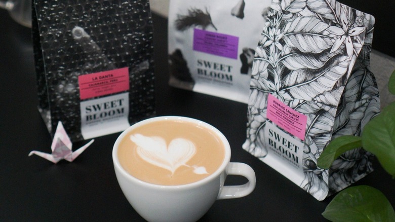 Cup of coffee with heart latte art next to bags of Sweet Bloom coffee