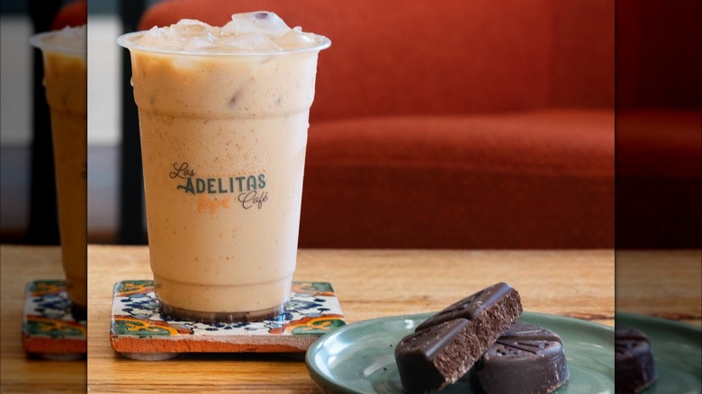 An iced coffee in a plastic Los Adelitas Café cub next to a plate of chocolate cookies.