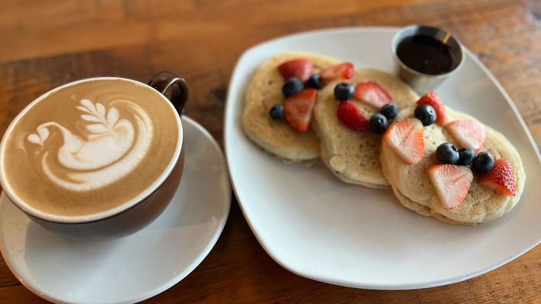 Cup of coffee with latte art next to a plate of small pancakes topped with fruit