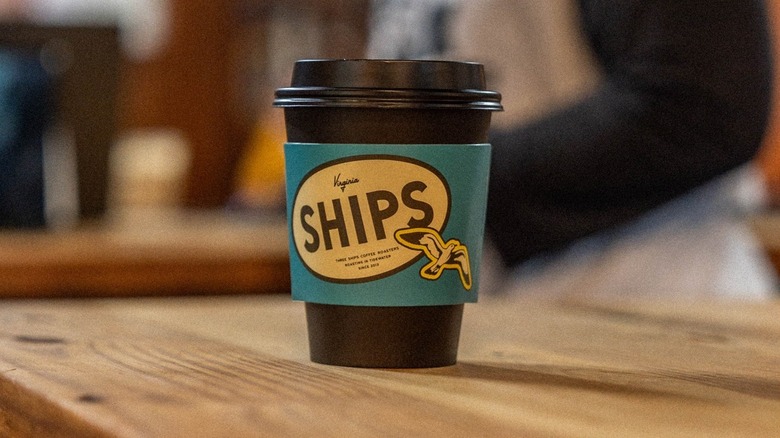 Black coffee cup with a green and brown sleeve that says "Ships" in large letters