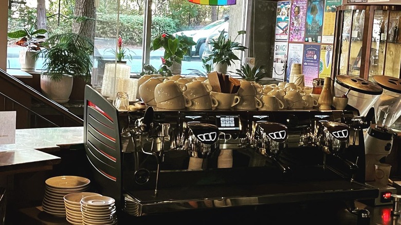 A silver espresso maker and stacked coffee mug sits on a café counter