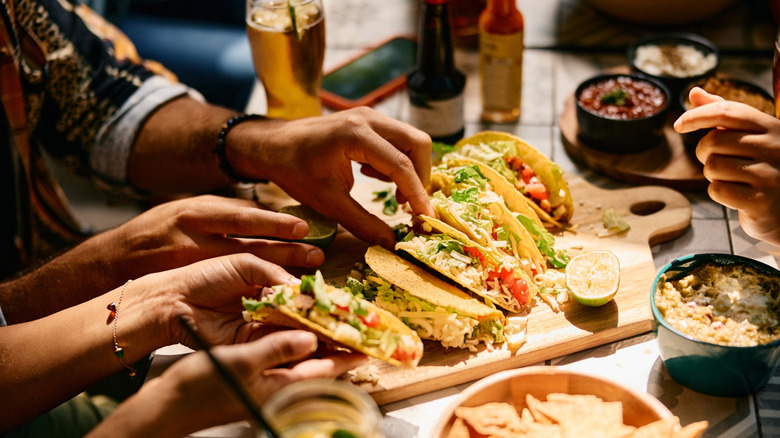 A wooden board of tacos surrounded by salsas on crowded table in Mexican restaurant