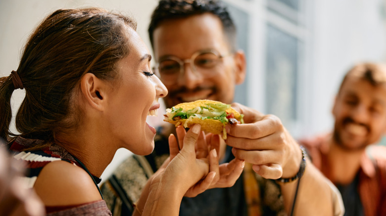 A smiling man holding a hard shell taco and holding it for a smiling woman to eat