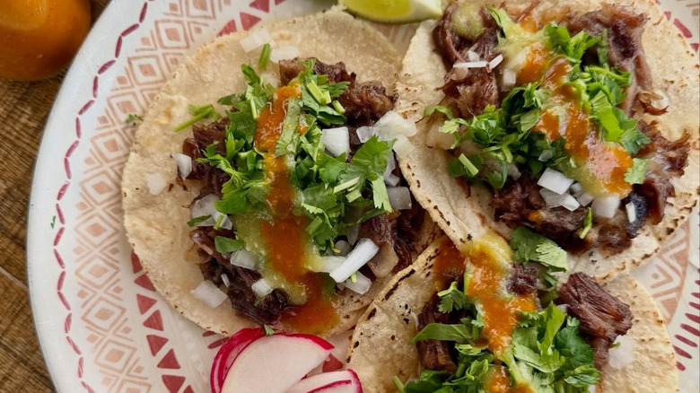 Three Mexican beef cheek tacos with toppings and lime wedges on plate