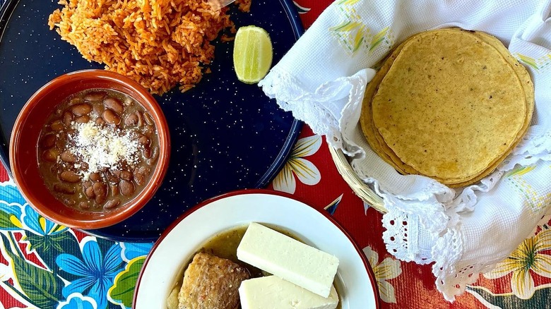 Pork belly in salsa verde with beans in dish, seasoned rice on plate, and basket of soft tortillas on decorative tablecloth