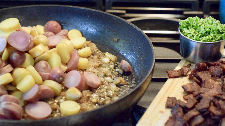 German potato salad being cooked ina skillet with bacon and parsley on the side sitting on a cutting board
