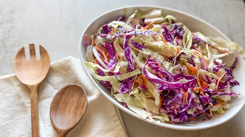 Coleslaw in a white bowl with two wooden serving utensils sitting on a napkin on the side