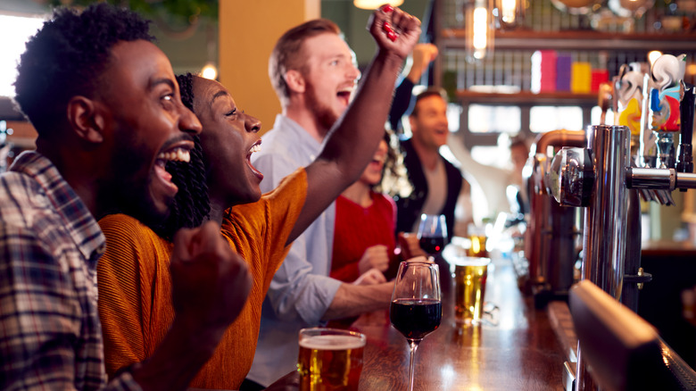 A crowd of people lined up at a bar counter, drinks in front of them, cheering. 