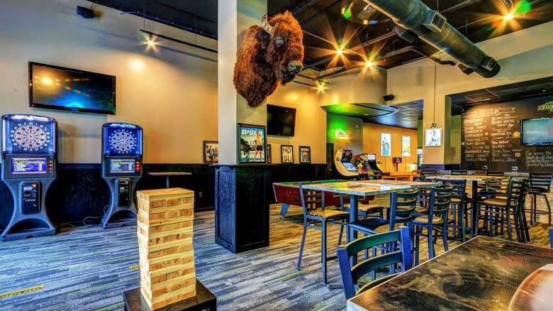 Room filled with tables, chairs, and arcade games, with a bison head mounted on a column. 