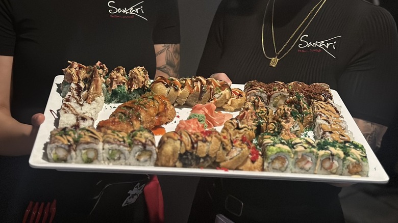Two people wearing shirts with Sakari's logo, holding a tray of assorted sushi rolls, regular ones as well as some deep-fried ones.
