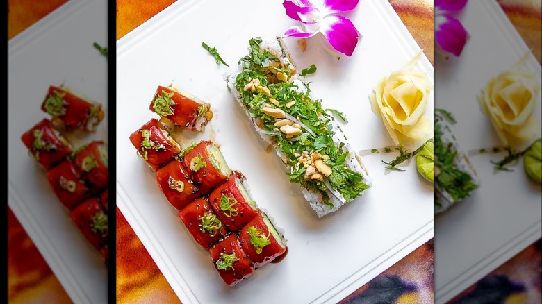 A tray with 2 different sushi rolls, one with salmon on top and the other one with nuts sprinkled on top, as well as a decorative flower on the right and ginger slices and wasabi