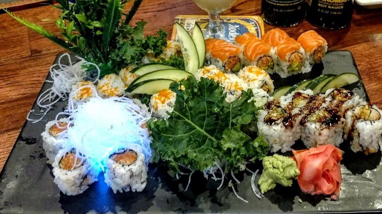 A tray with assorted sushi roll, with salmon, tuna, and avocado, alongside wasabi and ginger sclices, topped with lettuce and cucumber.