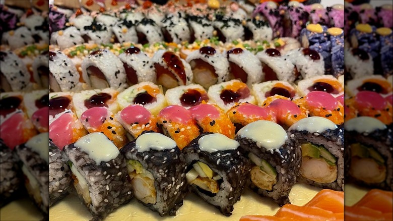 A tray of assorted sushi rolls, with black tint, others with purple tint and pink as well, as well as some others with salmon, and several different sauces on top