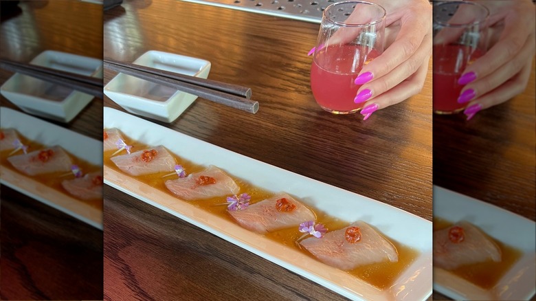 Raw fish on white tray next to hand holding pink drink